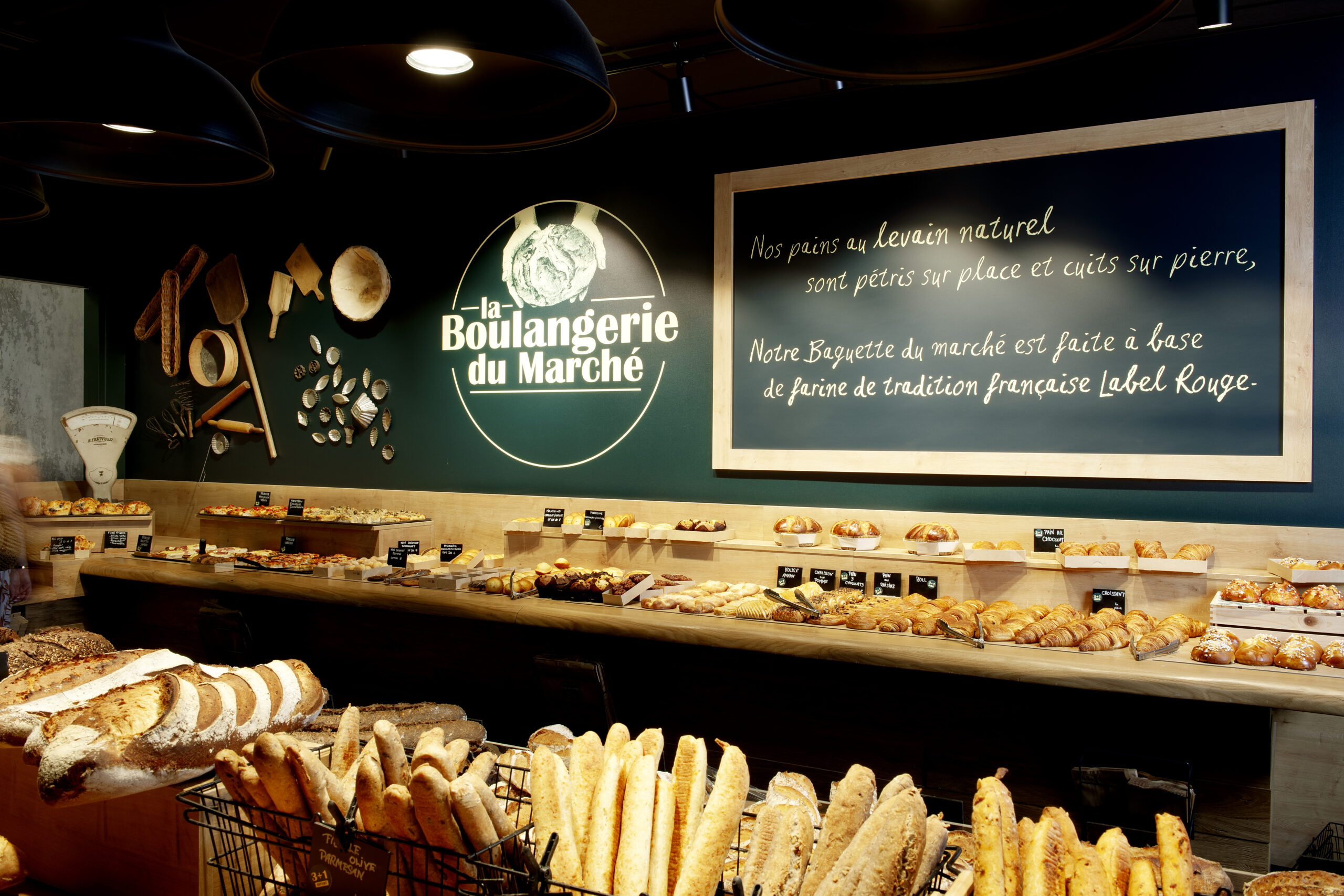 Cette image présente un étal généreusement garni de pains et de viennoiseries à la Boulangerie du Marché. En arrière-plan, une inscription met en avant les valeurs artisanales de la boulangerie, notamment l'utilisation de levain naturel et de farine Label Rouge. Les étagères en bois sont soigneusement organisées, avec des produits tels que des baguettes, croissants, et autres douceurs traditionnelles françaises. L'éclairage doux et la décoration traditionnelle renforcent l'ambiance chaleureuse et authentique de l'établissement.