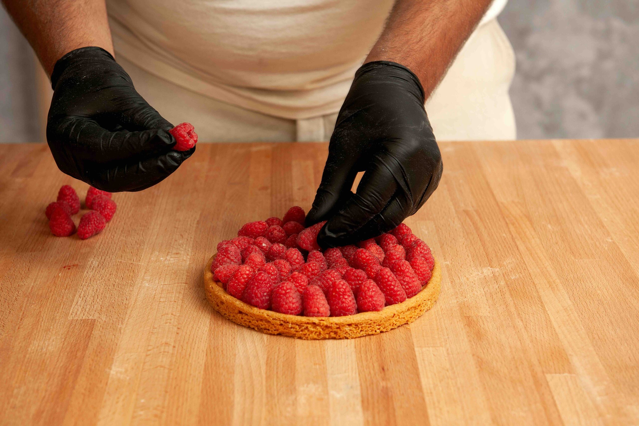 L'image montre un pâtissier en train de disposer délicatement des framboises fraîches sur une base de tarte dorée. P