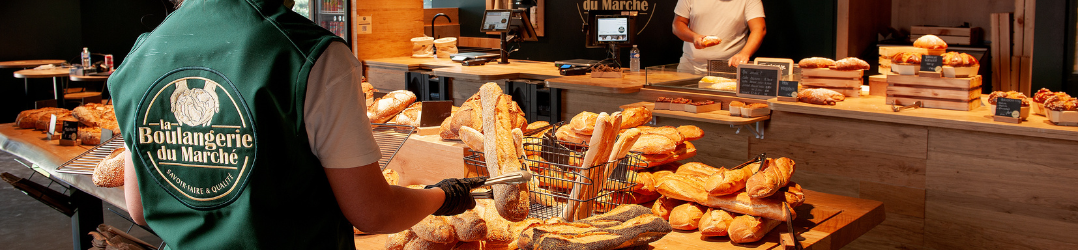 L'image montre un employé de la Boulangerie du Marché en train de préparer une commande de baguettes fraîches. Le logo de la boulangerie est visible sur son uniforme, soulignant l'identité de la marque. À l'arrière-plan, le comptoir est rempli de pains et de viennoiseries soigneusement disposés, tandis qu'un autre employé sert les clients à la caisse. L'atmosphère est chaleureuse et artisanale, avec une attention particulière portée à la présentation des produits et au service client.